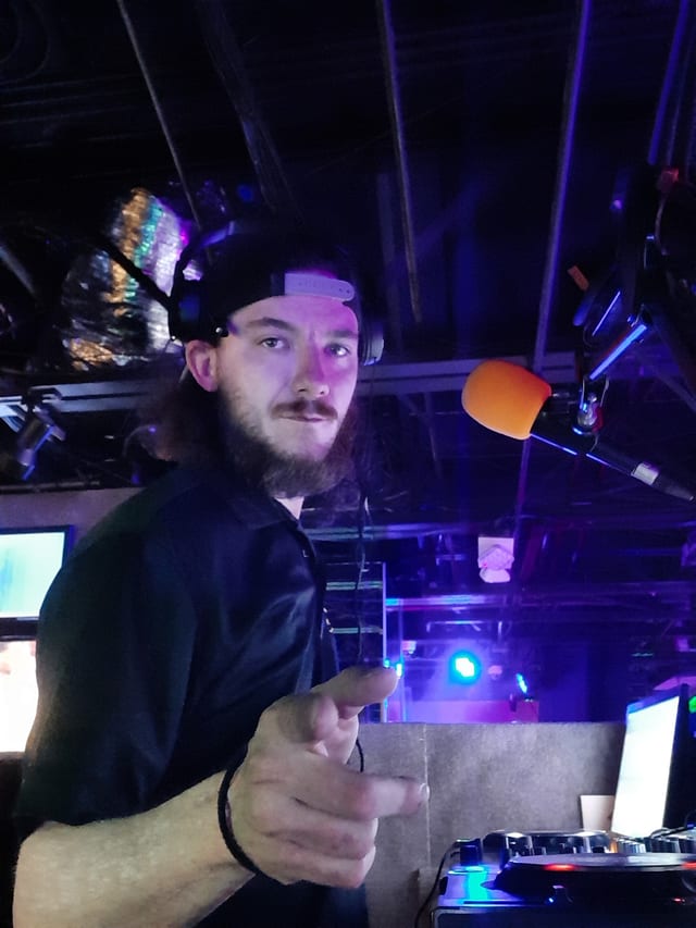 Andy in the DJ booth, headphones on, pointing at the camera