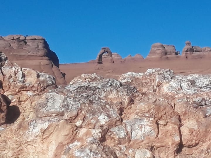 Delicate Arch seen far off on a ridge of red sandstone above a foreground of weathered rock
