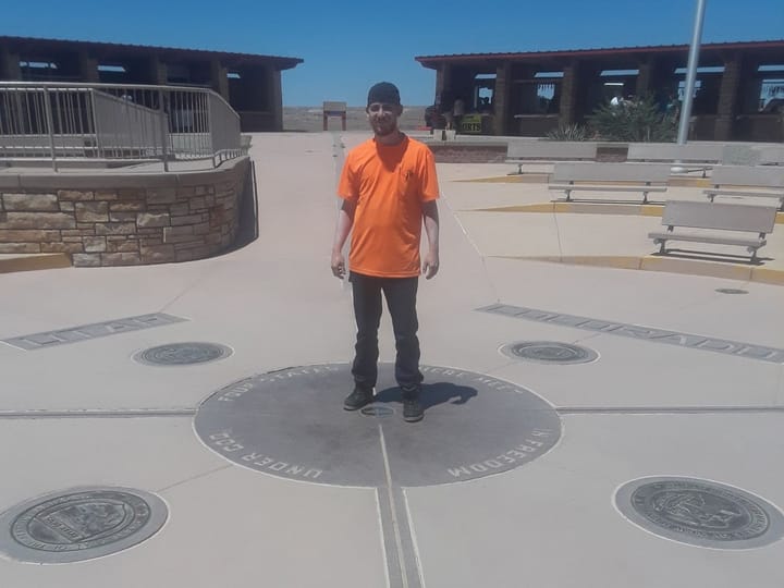 Standing on the Four Corners marker in an orange shirt, with the Utah and Colorado plaques in the pavement