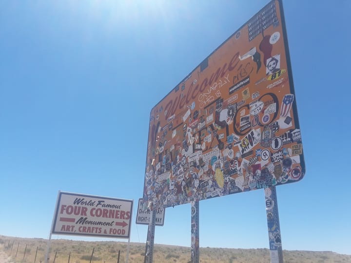 A New Mexico welcome sign covered in stickers beside a Four Corners Monument sign
