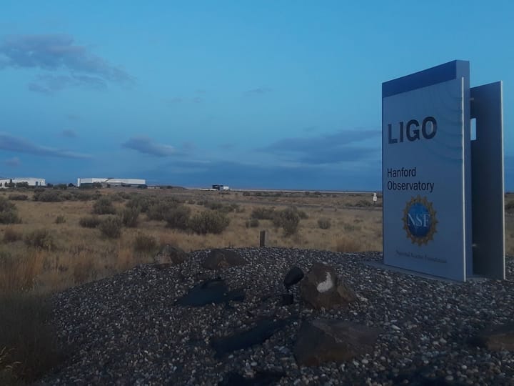 The LIGO sign at dusk with the observatory buildings on the horizon
