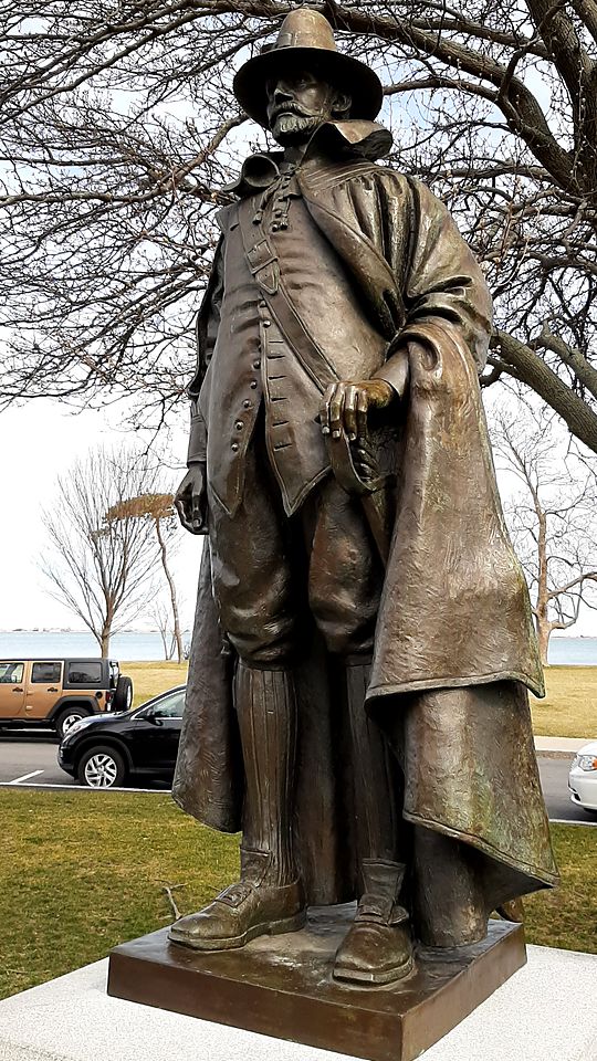 A bronze statue of a Pilgrim in a wide-brimmed hat and cloak, with the waterfront behind it
