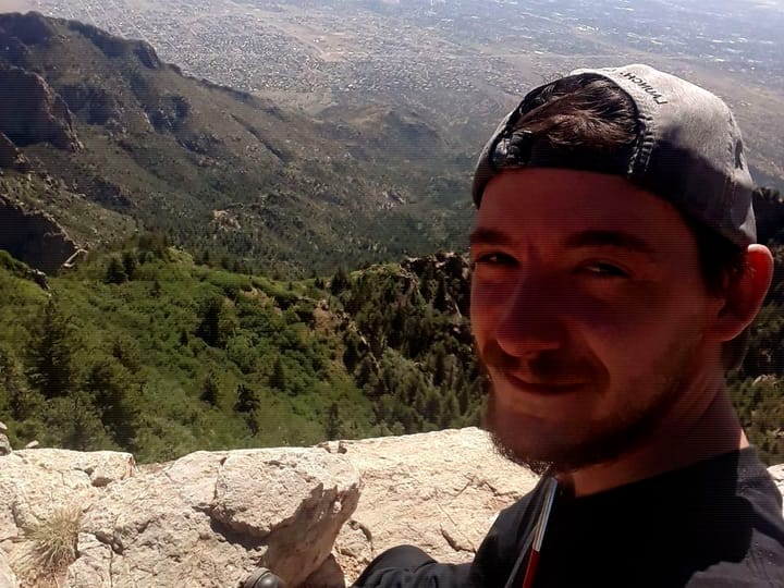 A selfie in a backwards cap at a stone overlook, with forested slopes and the city of Albuquerque far below