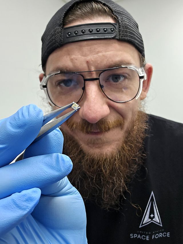 Andy in safety glasses and blue nitrile gloves, holding a tiny component up to the camera with tweezers